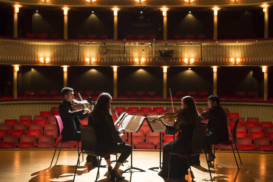A string quartet, wearing smart outfits, performing together on stage, in front of an theatre.
